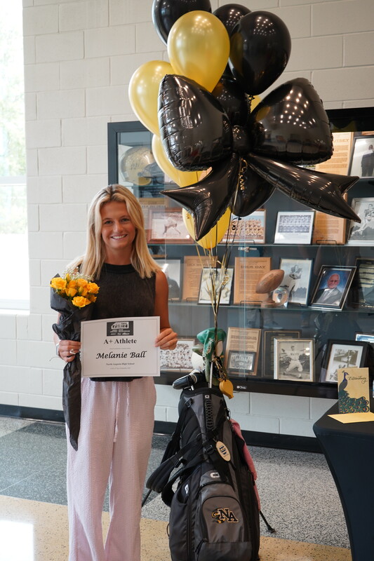 Student standing in front of trophy case with a set of golf clubs, balloons, yellow flowers, and award certificate.