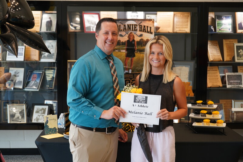 Student and principal standing in front of trophy case holding a certificate and flowers.