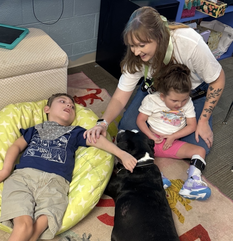 A boy is laying back on a beanbag with his hand on a black and white dog with the help of teacher, Ms. Bostedor. A girl is sitting next to the dog.