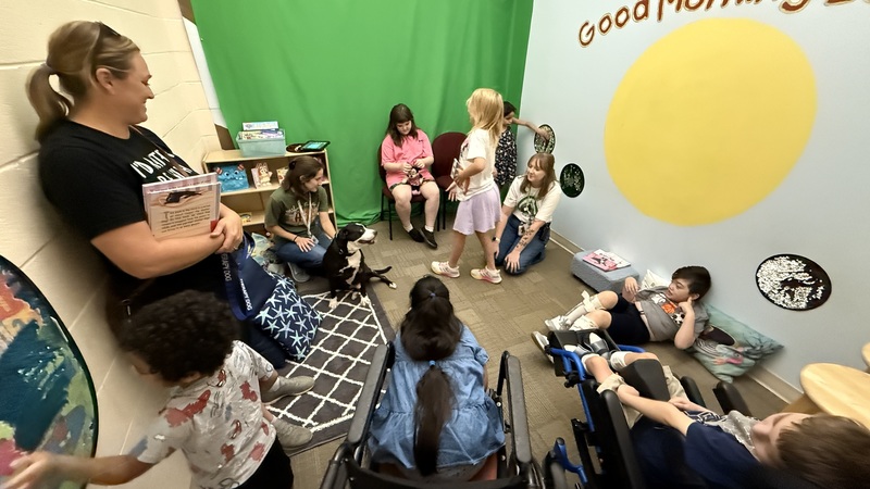 A seven students and three adults are gathered together around a black and white dog that is sitting on the floor.