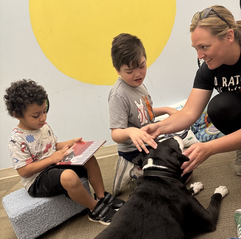 One boy is seated and holding a book. Another boy is being helped by an adult to pet a black and white dog.