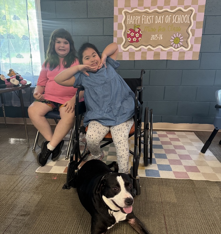 Two girls, one in a chair, the other in a wheelchair, sit behind Cooper, a black and white dog.