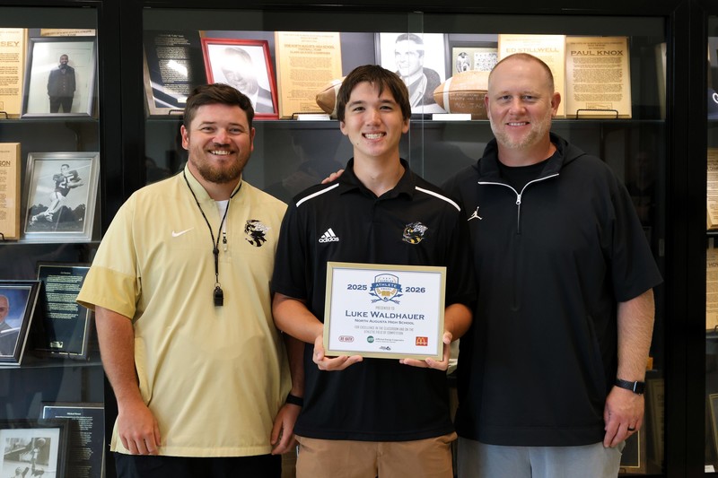 2 Coaches on either side of student holding award in front of trophy case. 