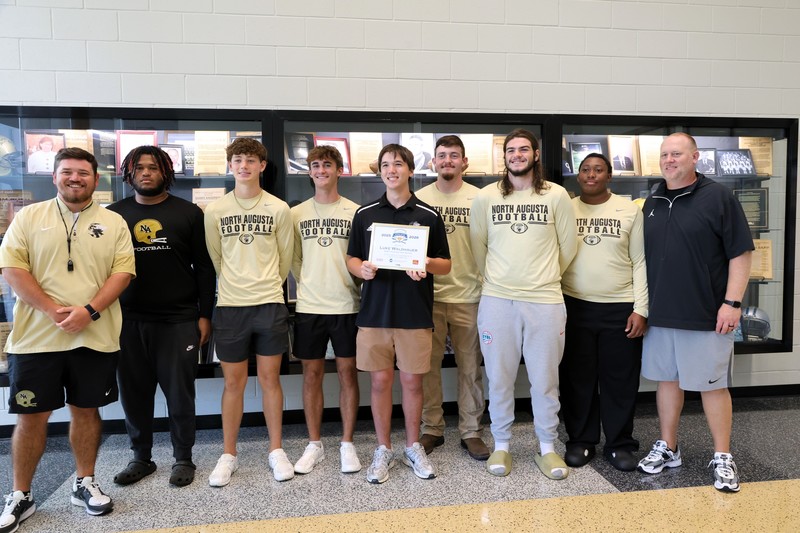Football Players surrounding student with award in front of trophy case. 