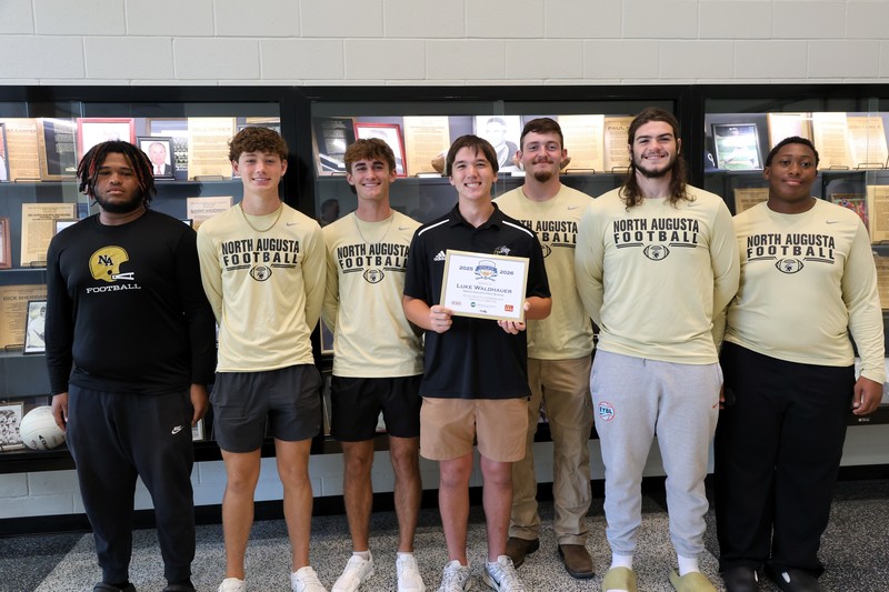 Football Players surrounding student with award in front of trophy case. 