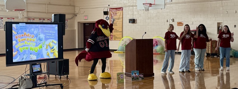 A group of students seated on bleachers watch a presentation by Cocky's ReadingUSC college students, along with the mascot, Cocky., stand in front of a screen that projects a book.