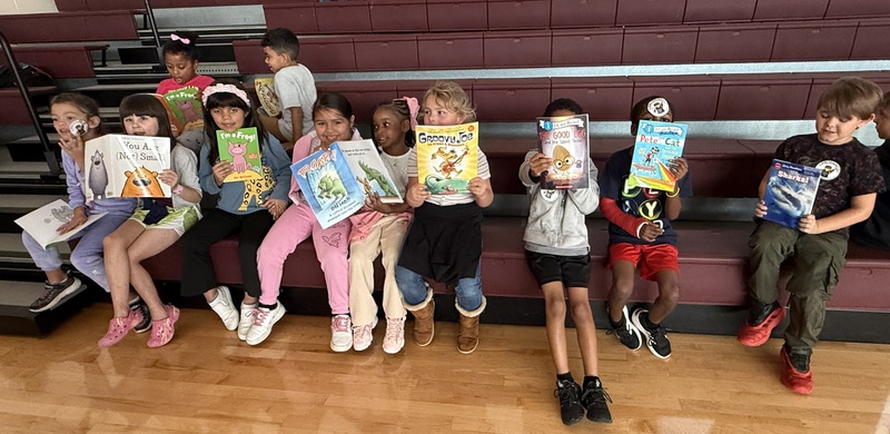 A group of students seated on a bleacher hold up books.