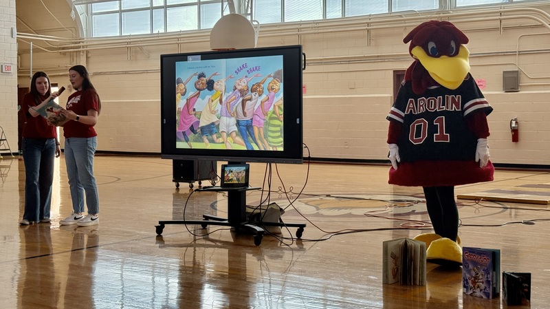 Cocky and two college students read from a book, which is projected on a screen.