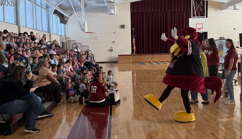 A group of students seated on bleachers watch a presentation by Cocky's Reading Express by USC college students along with the mascot, Cocky.