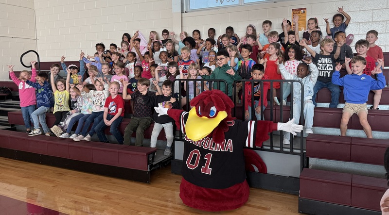 A group of excited students seated on bleachers with the USC mascot posing in front of them.