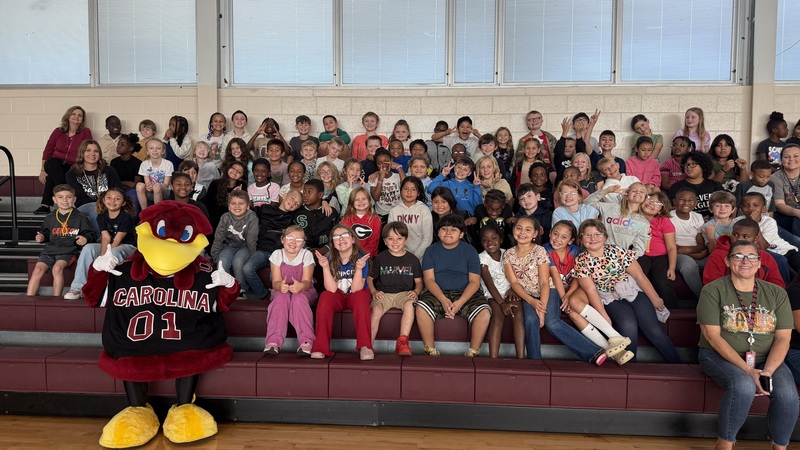 Third grade students and teachers are seated on bleachers along with Cocky, the USC mascot.