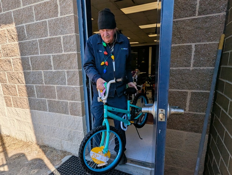 A man wearing holiday lights around his neck is walking a bike out of a door.