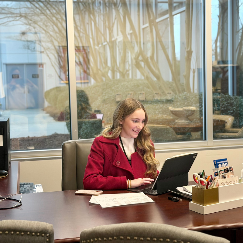 Student in a red coat sitting at a desk working on a computer.