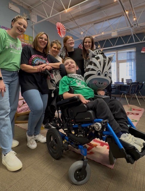 Five teachers stand behind a boy in a wheelchair.