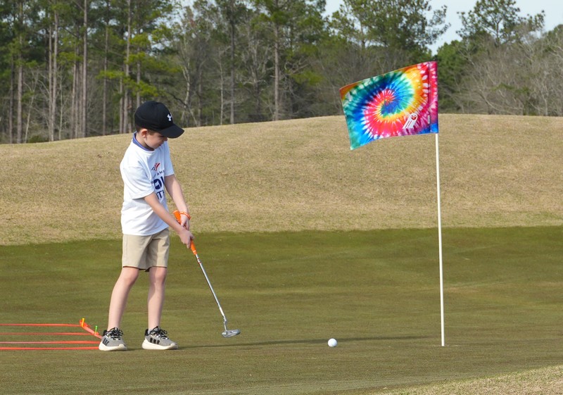 Kid putts golf ball towards tie dye flag on golf course.