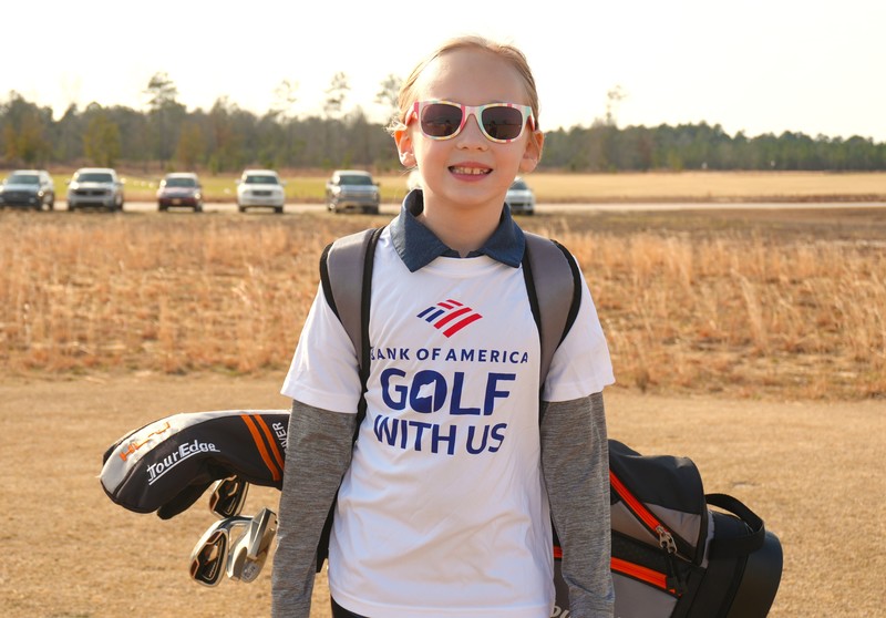 Kid poses with Bank of America Golf with Us Shirt and golf bag.