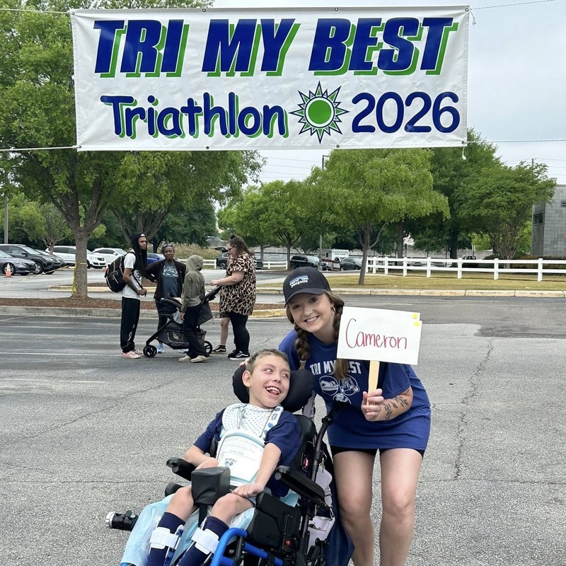 A teacher holding a sign stands next to a smiling boy in a wheelchair outside.