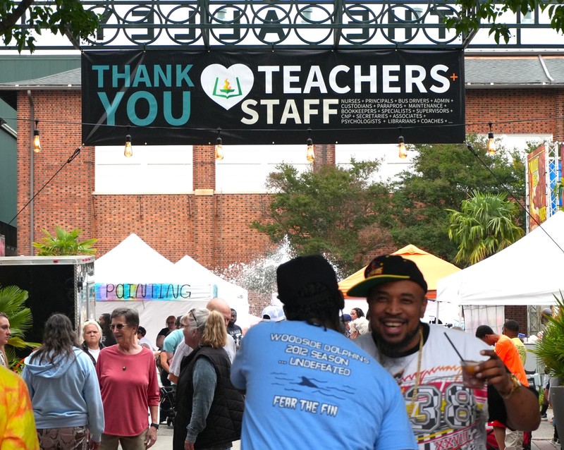 Two men hug beneath a banner at a festival that reads "Thank You Teachers and Staff."