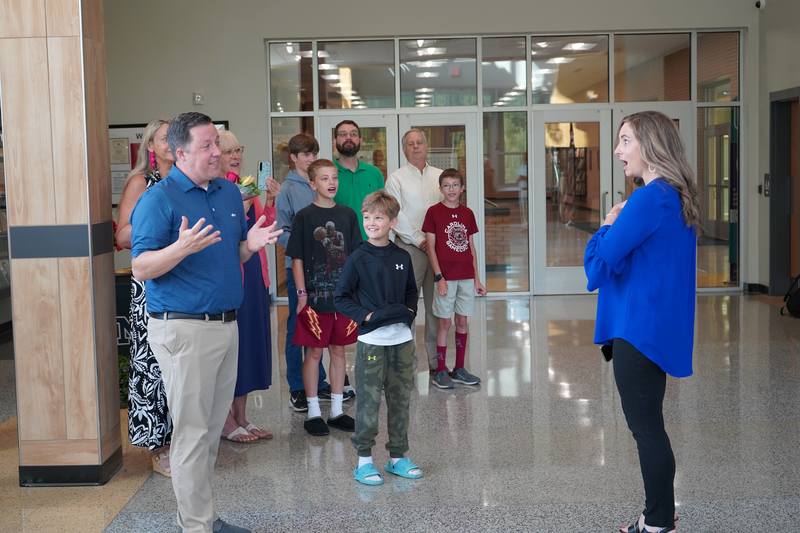 Teacher Surprised by Principal with family looking on in school lobby.