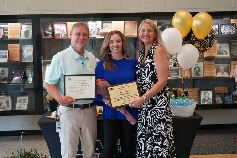 Winner with plaques and two counselors on either side with trophy display case in background and balloons on table. 