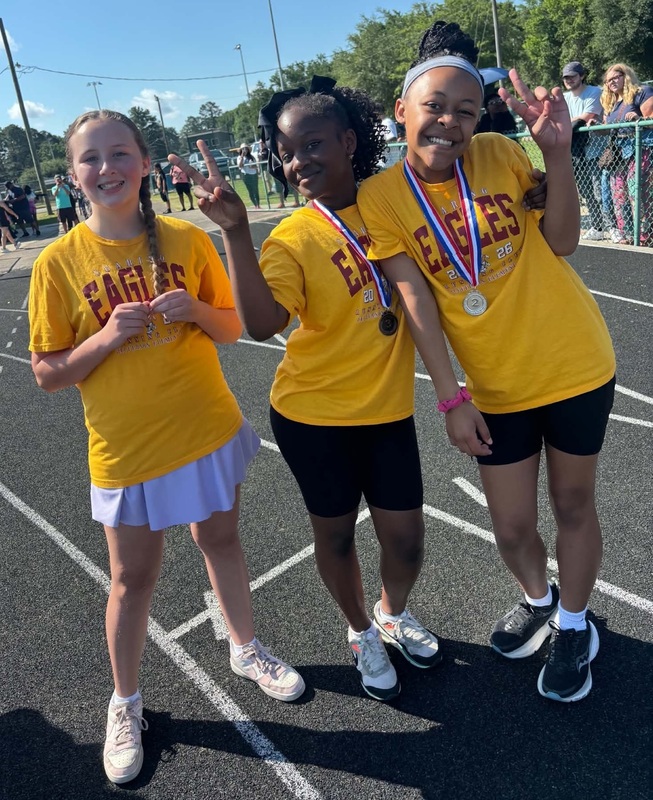 Three girls, two wearing medals, stand side-by-side.