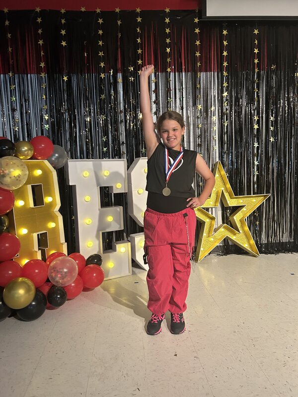Girl with medal standing in front of RES sign with one arm up.
