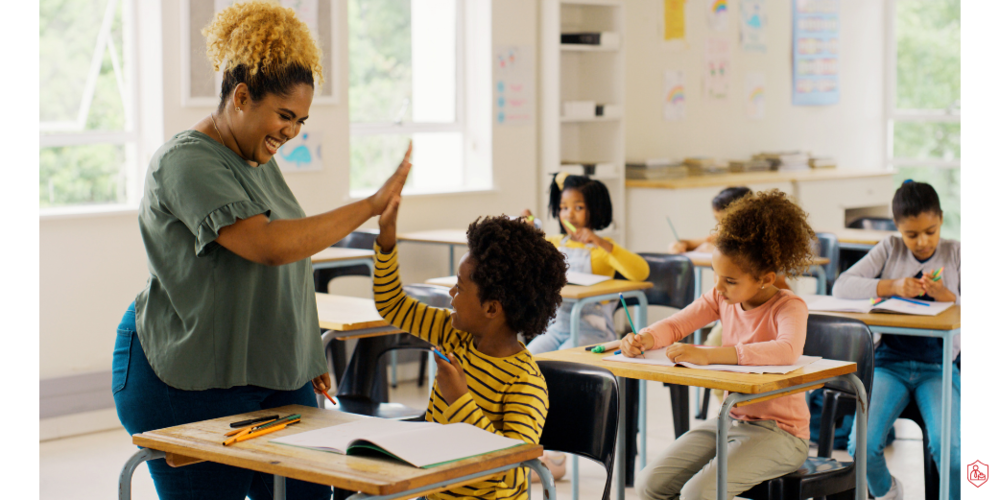 TEACHER  HIGH-FIVING A STUDENTE WITH OTHER STUDENTS IN DESKS BEHIND THEM