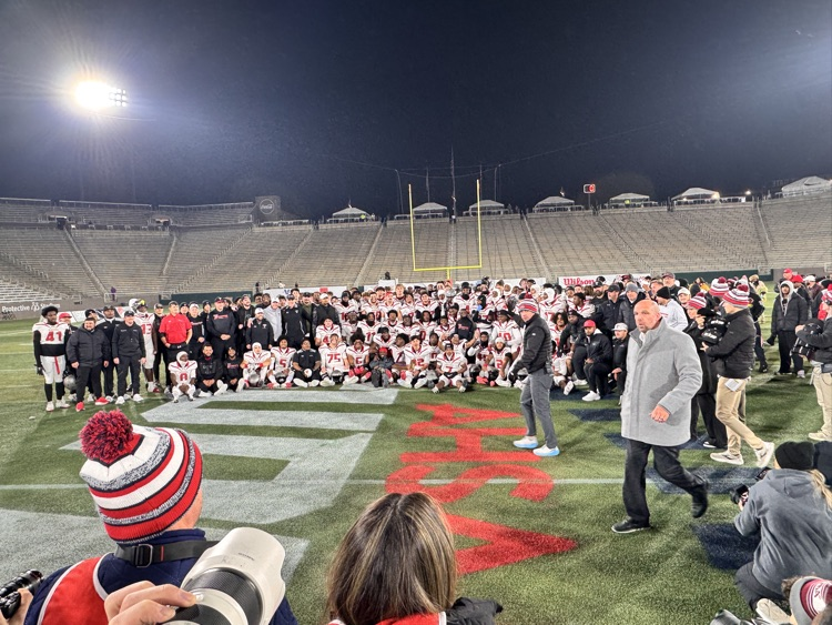 wide shot of lots of football players and spectators, watching them pose for a photo with lots of camera lenses pointed at them