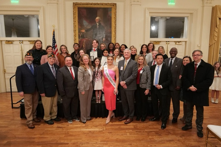 large group of people standing in a ballroom with a wooden floor and a large portrait of George Washington on the wall and an American flag on the left side of the photo