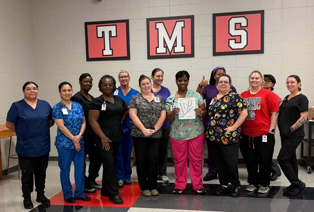 13 cafeteria personnel gathered; center one holding health inspection sheet with large "100" written on it