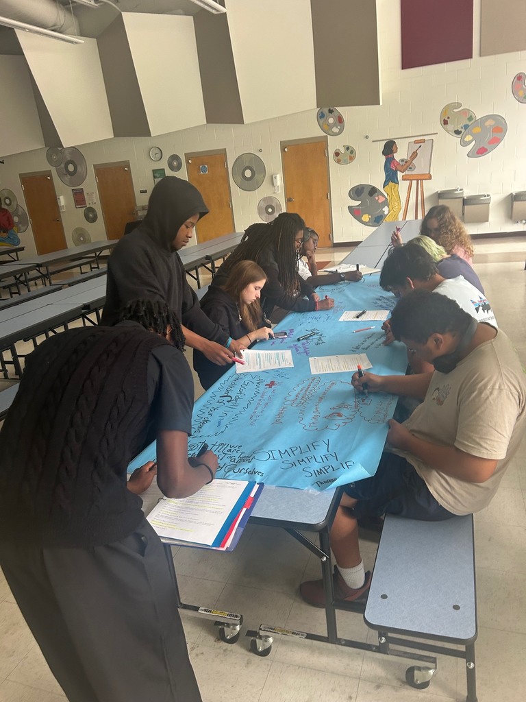 Many students sit as one stands to work on project together