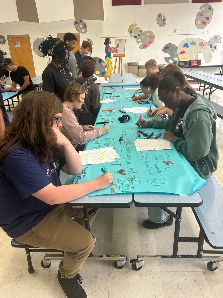 3 students stand as several others sit and create drawing on bulletin board paper