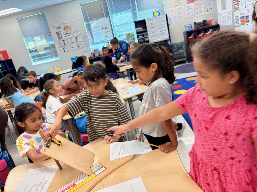 students working at tables with clipboards, toy cars, and rulers