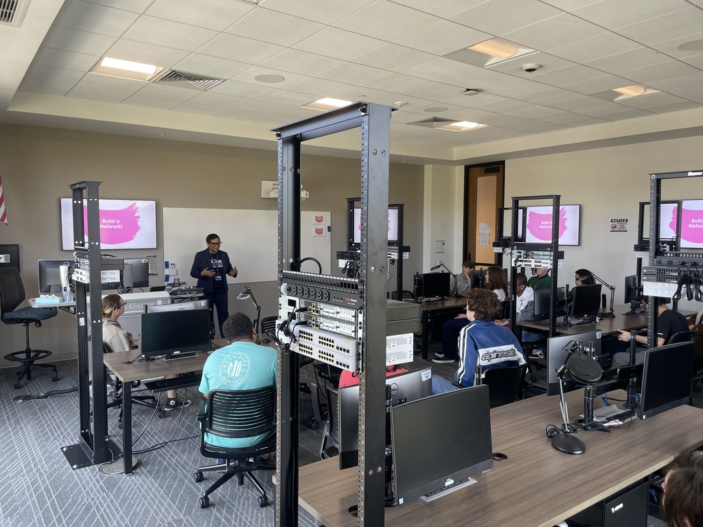 students sit in a room filled with computers as teacher speaks at front of the room