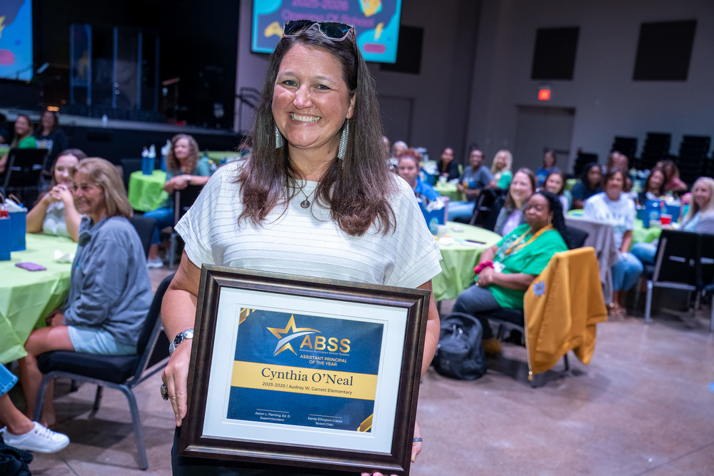 Cynthia O'Neal holding a Assistant Principal of the Year plaque in the middle of a group of staff sitting at tables