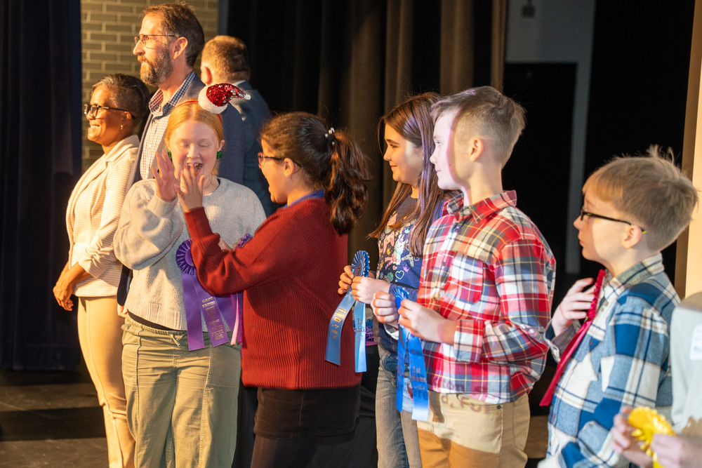 Students stand on stage with recognition ribbons at a Science Fair ceremony.  Two girls high five one another.