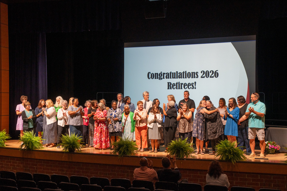 A large group of retirees on stage at a retirement celebration with a large presentation slide behind them that says Congratulations 2026 Retirees!