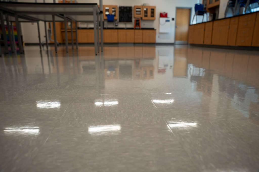 A low-angle view of an extremely polished classroom floor that creates perfect reflections of the tables and chairs above. The high-gloss finish showcases the quality of the custodial team's work.