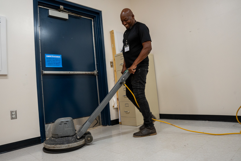 A smiling custodial worker operates a floor buffer in what appears to be an office area, with blue doors and filing cabinets visible in the background. His expression shows pride in the meticulous work being performed.