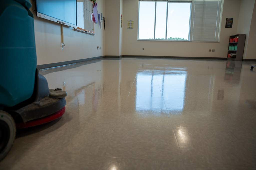 A pristine classroom with spotless, reflective floors that mirror the windows and ceiling lights. The room features white walls, large windows, and appears freshly cleaned and ready for students.