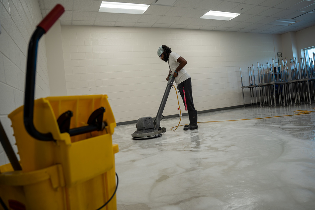 A worker operates a large floor buffing machine in what appears to be a school cafeteria or multipurpose room. Yellow cleaning equipment is visible in the foreground, and the worker is focused on polishing the floors to a high shine.