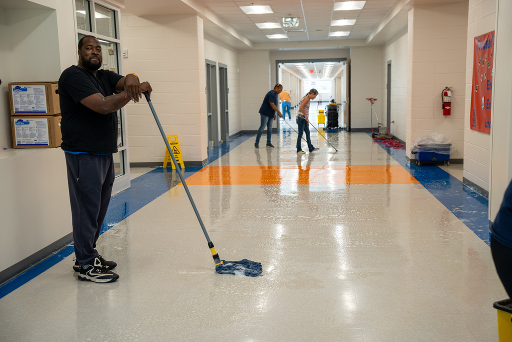 A custodial worker in a dark shirt and jeans stands smiling in a school hallway, holding a mop. Behind him, other workers are cleaning the colorful tiled floor with professional equipment. The hallway features white walls, large windows, and a vibrant orange and blue geometric pattern on the floor.