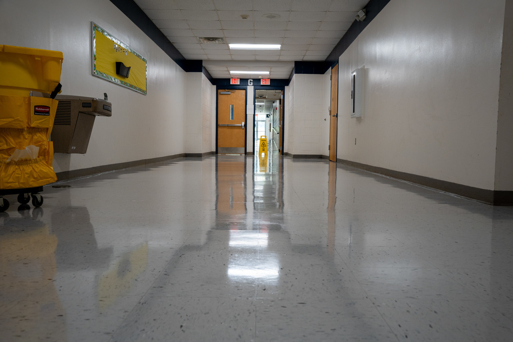A perfectly polished school hallway stretches into the distance, with floors so clean they create mirror-like reflections of the ceiling lights and exit signs. Yellow cleaning equipment sits against the wall, evidence of the thorough work completed.
