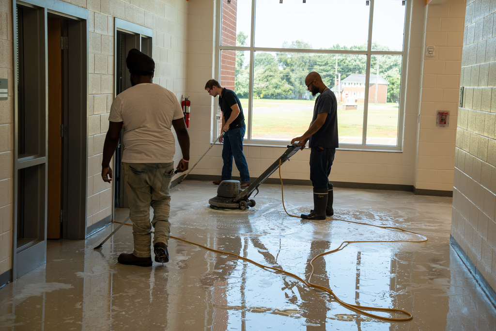 Multiple custodial workers clean a school hallway using professional floor equipment and hoses. The team works together with industrial cleaning machines, demonstrating the thorough deep-cleaning process taking place throughout the summer.