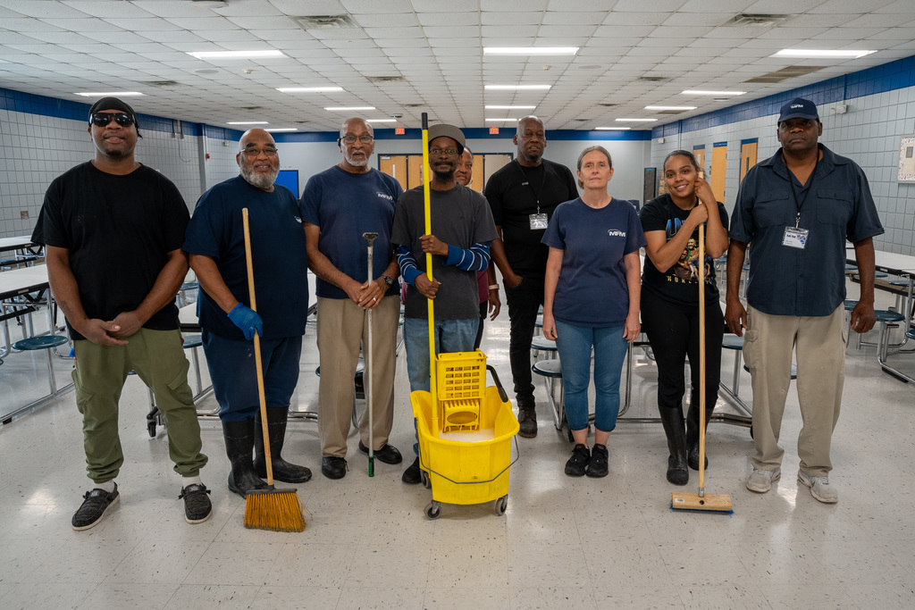 A large group of custodial workers and administrators pose together in a school cafeteria, holding various cleaning tools including mops, brooms, and a yellow mop bucket. The diverse team displays the collaborative effort behind the school's summer deep-cleaning initiative.