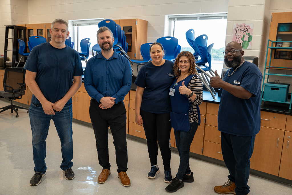 A group of five people, including custodial staff and school administrators, pose together in a classroom with stacked blue chairs visible in the background. The floors shine brilliantly, reflecting the thorough cleaning work completed.