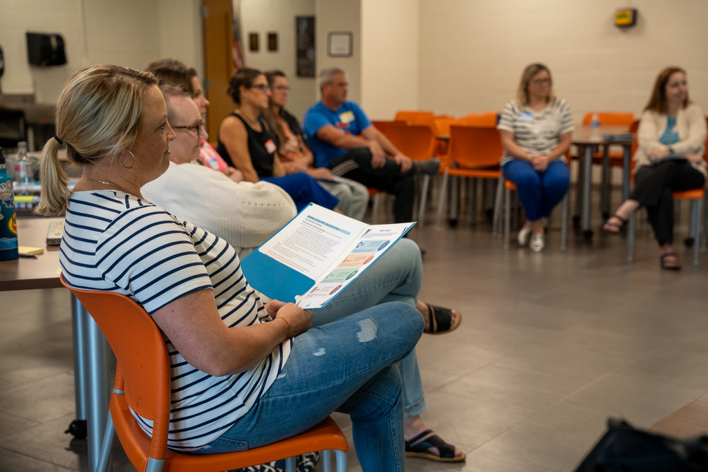 A room of around 10 educators sitting in a semi-circle, the one in the foreground has training materials in a folder open on her lap