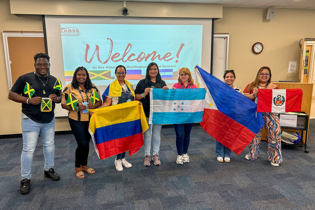 Seven Participate Learning Ambassador Teachers stand in a row holding flags from their home countries in a classroom setting. From left to right, they hold flags of Jamaica (green, yellow, and black), Colombia (yellow, blue, and red), Honduras (blue and white with stars), Philippines (blue, white, and red with yellow sun and stars), and Peru (red and white with coat of arms). Behind them is a projection screen displaying "Welcome! to the Alamance-Burlington School System" with colorful flag graphics. The teachers are smiling and wearing casual clothing with name badges, standing on gray carpeting in what appears to be a school conference or training room.