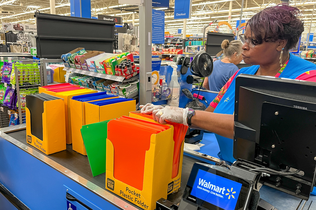 Walmart cashier in blue vest and gloves scanning colorful pocket folders and school supplies at checkout register, with various educational items visible on counter.