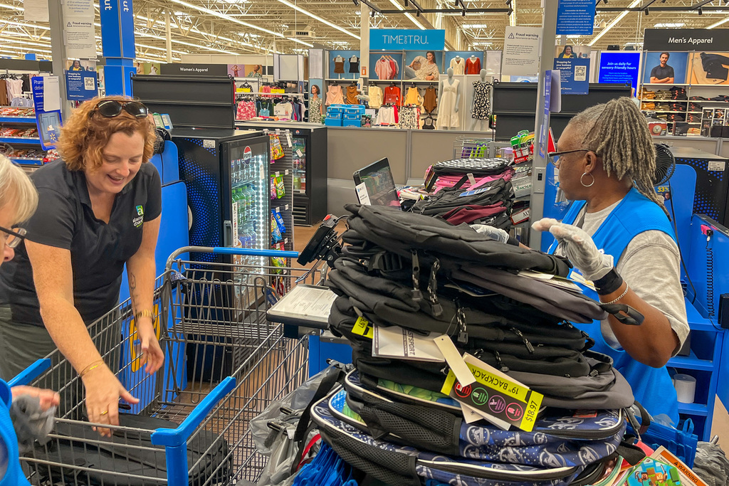 Woman in dark shirt loading school supplies into shopping cart while Walmart employee in blue vest and gloves helps organize backpacks at checkout counter inside store.